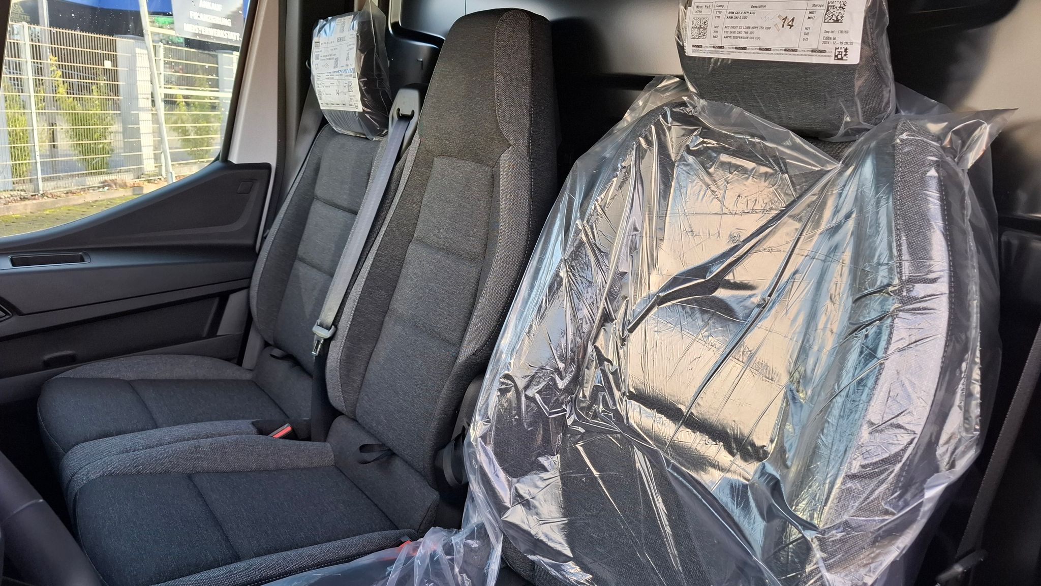 Grauer Stoff-Fahrzeugsitz mit Sicherheitsgurt in modernem Autohaus-Showroom mit spiegelndem Fliesenboden und Loungebereich im Hintergrund