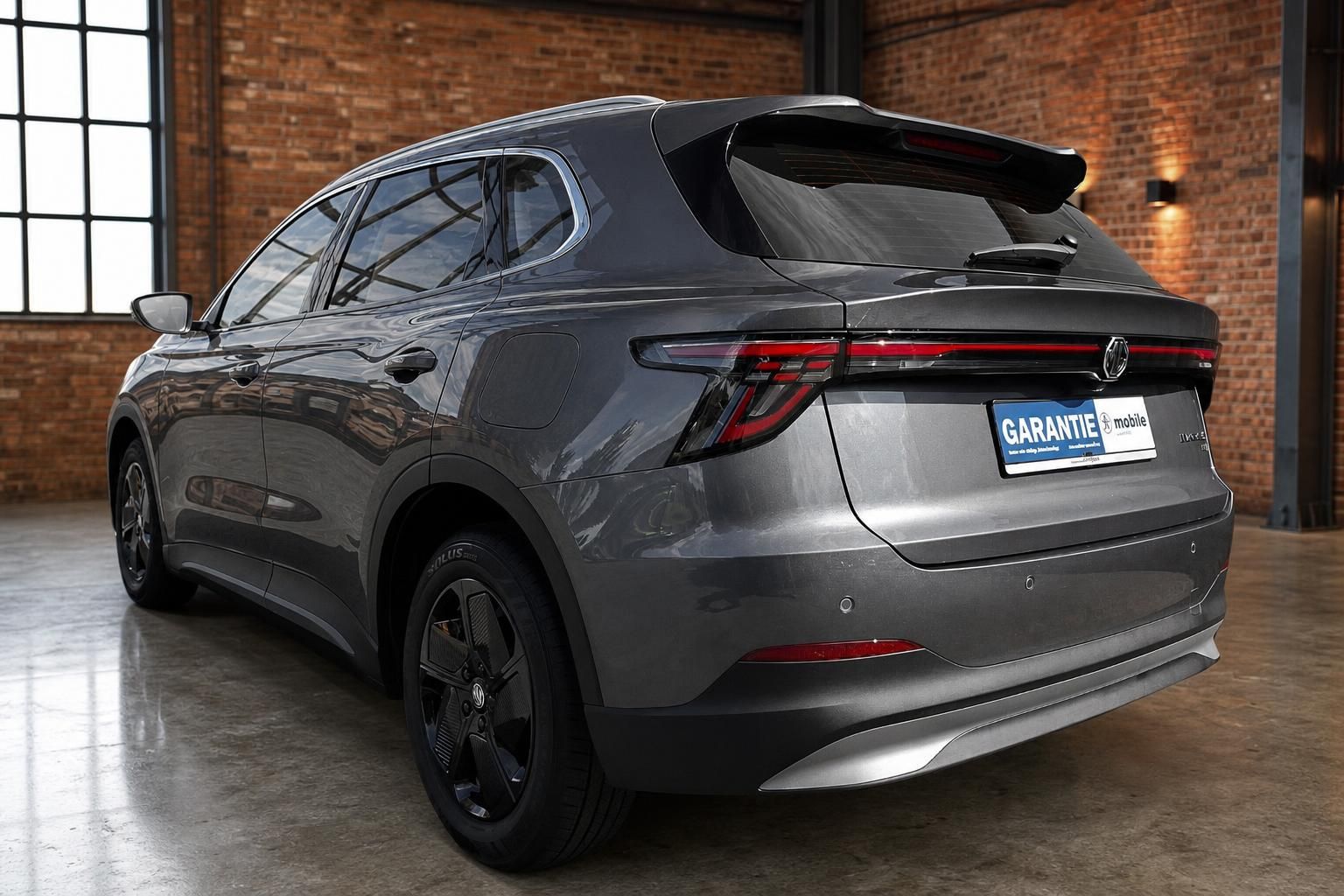 MG SUV Elektro Grau, Heckansicht schraeg mit Dachspoiler in Fabrikhalle