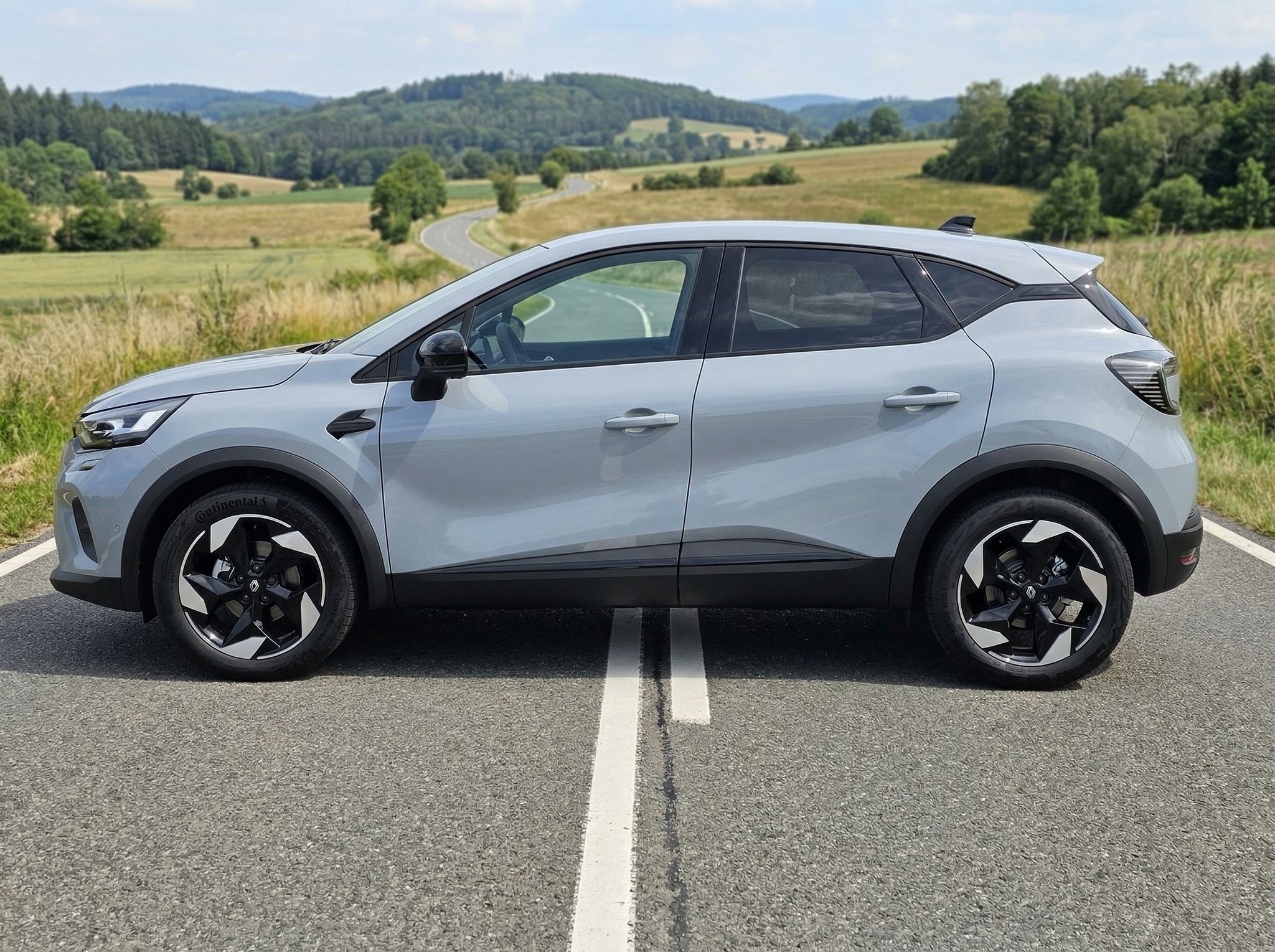 Renault Captur silber SUV Seitenansicht auf Asphalt vor ländlicher Kulisse mit Feldern und Hügeln