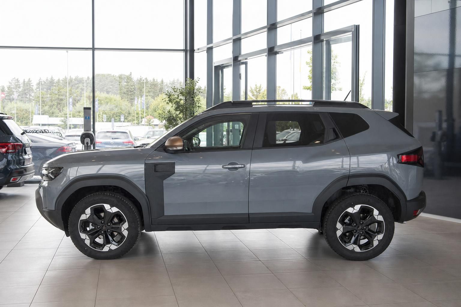 Grauer Dacia Duster SUV - komplette Seitenansicht von links im Autohaus mit Panoramafenstern