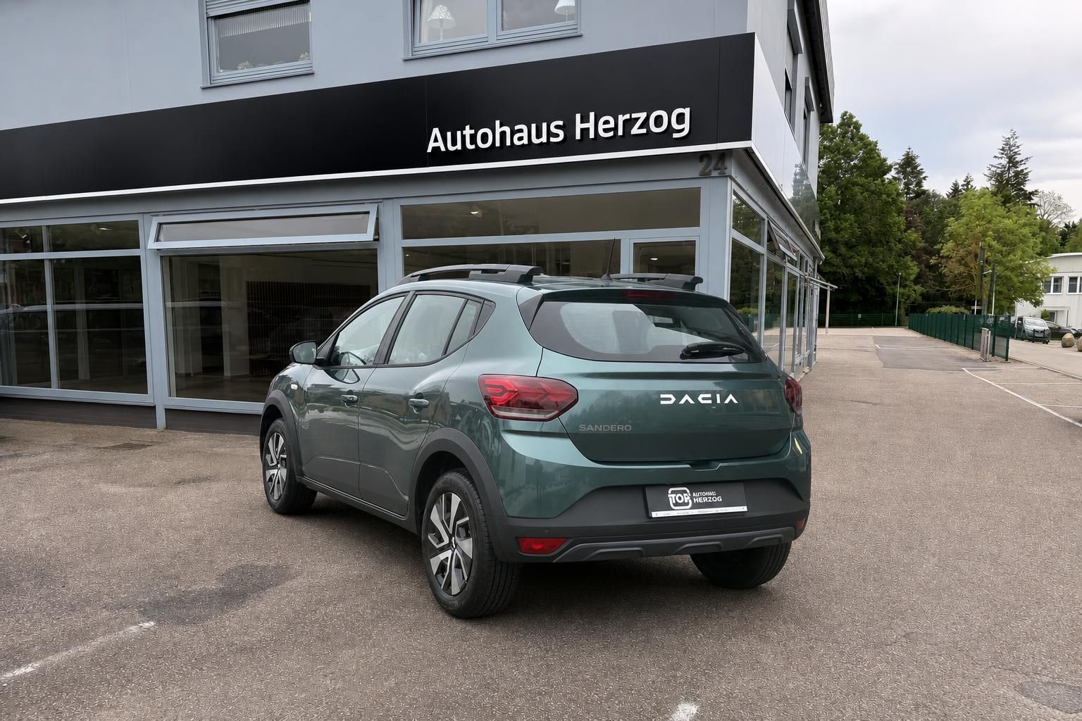 Dacia Sandero grün metallic Heckansicht mit Autohaus Herzog im Hintergrund