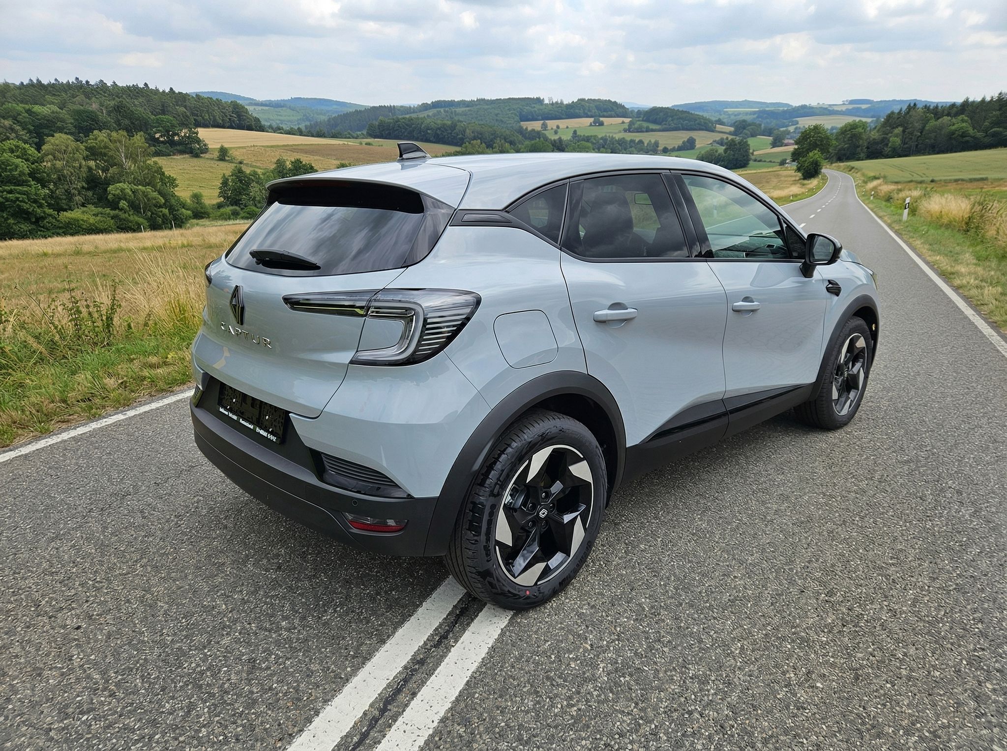 Renault Captur SUV silber Heck-Seitenansicht auf Straße mit Feldern und Wäldern